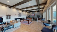 This single-story community clubroom in Humble, Texas, is thoughtfully designed to blend social functionality with refined aesthetics. The expansive open-plan layout is anchored by striking dark wood coffered beams overhead, recessed lighting, and warm light wood flooring that flows throughout the entire space.

The central gathering area features a full-sized pool table atop a richly colored area rug, flanked by comfortable lounge seating. Adjacent to this game zone, a dedicated lounge nook showcases bold decorative wallpaper in an art deco-inspired pattern, mounted flat-screen televisions, and curated furniture arranged for relaxed viewing and conversation.

The kitchen bar area is a standout feature, finished with deep-toned granite countertops, dark wood cabinetry, and a shimmering stacked tile backsplash that extends floor to ceiling. Copper pendant lights add warmth above the counter, complementing open wooden shelving stocked for entertaining. A beverage station, sink, and under-counter refrigeration complete this well-appointed area.

Large glass French doors along the perimeter flood the space with natural light and provide direct access to an outdoor pool area, seamlessly connecting interior and exterior living. The overall ambiance is upscale yet inviting, ideal for community gatherings.