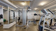 Located at 640 Main St in Saskatoon's Nutana neighbourhood, this fitness facility at Nutana Towers offers a compact yet comprehensive exercise space across a single floor. The layout includes a dedicated exercise room and a connecting hallway, making efficient use of the available area.

The exercise room is well-appointed with a diverse range of equipment to support both cardiovascular and strength training. Cardio machines include a treadmill, stationary bike, and elliptical trainer, while strength training is facilitated by a multi-station cable machine, adjustable benches, a weight rack, and free weights including barbells and plates. A mounted flat-screen television adds convenience for users during workouts.

The room features durable speckled rubber flooring suited for high-impact activity, and full-length mirrored walls along one side that visually expand the space and assist with exercise form. The exposed ceiling with visible ductwork and evenly spaced flush-mount lighting gives the room a utilitarian, functional aesthetic. Small windows along the exterior wall allow for a modest amount of natural light.

The adjacent hallway provides a clean transition into the facility, finished with the same neutral grey tones and smooth flooring. Informational signage is posted near the entrance, and a glass-panelled door contributes to an open, accessible feel. Overall, the space is practical, clean, and purpose-built for fitness.