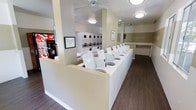 This single-floor community laundry facility is designed for efficiency and convenience. The space is organized around a central dividing counter that neatly separates the room into functional zones. A long row of top-loading washing machines lines one side, while a bank of stacked dryer units occupies the back wall, providing ample laundry capacity for multiple users at once. The flooring throughout is a dark wood-look surface that contrasts cleanly with the bright white appliances and light-toned walls. Ceiling-mounted flush lights and a ceiling fan ensure the space remains well-lit and well-ventilated. A Coca-Cola vending machine is positioned near the entrance, adding a convenient amenity for visitors during their stay. Informational signage is posted on the walls, keeping users informed of guidelines and instructions. A folding or sorting counter is visible along one wall, offering a practical surface for managing laundry. The word "CLEAN" is displayed prominently on a mirrored or accent wall panel, reinforcing the facility's tidy and well-maintained atmosphere. Overall, the space is thoughtfully laid out to accommodate high usage while maintaining a clean, orderly environment.