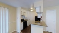 This single-story space features a well-organized kitchen as its central highlight. The kitchen is outfitted with white upper and lower cabinetry that provides ample storage, complemented by granite-look countertops that run along the cooking area and extend to a built-in peninsula. The peninsula includes a stainless steel sink, offering a practical workspace that also serves as a natural divider between the kitchen and the adjoining living area. Black appliances, including a gas range with an overhead ventilation hood, anchor the cooking zone and contrast cleanly against the white cabinetry. The flooring transitions from dark hardwood-style material in the kitchen to carpet in the adjacent living space, subtly defining each zone. Ceiling-mounted flush lighting provides consistent illumination throughout. The walls are painted in a neutral warm tone, contributing to a clean and bright atmosphere. A fire extinguisher is visibly mounted near the kitchen entrance, and a door leads to an additional area beyond the cooking space, suggesting further functional rooms within the unit.