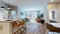 This single-floor residence offers a thoughtfully designed layout that balances open communal spaces with private retreats. The living area serves as the heart of the unit, featuring warm wood-look flooring, soft light-blue walls, and a large sliding glass door that floods the room with natural light and provides access to an outdoor balcony. A gallery wall adds character, while the generous layout easily accommodates a sectional sofa, accent seating, and a bar cart near the kitchen transition.

The kitchen is well-appointed with sleek white cabinetry, stainless steel appliances, subway tile backsplash, stone-topped counters, and pendant lighting overhead. Wood-look flooring continues throughout, complementing the clean, modern aesthetic.

The unit includes two bedrooms. One is carpeted and features a desk area, a wall-mounted television, and a lounge chair with ample natural light. The other offers a similar neutral palette with direct sightlines to the kitchen. A spacious walk-in closet provides generous storage. The bathroom is finished with light tile flooring, an extended double-sink vanity with abundant drawer storage, a large mirror, and a shower with a decorative curtain, creating a clean and functional space.