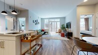 This single-floor residence offers a thoughtfully designed layout that balances open communal spaces with private retreats. The living area serves as the heart of the unit, featuring warm wood-look flooring, soft light-blue walls, and a large sliding glass door that floods the room with natural light and provides access to an outdoor balcony. A gallery wall adds character, while the generous layout easily accommodates a sectional sofa, accent seating, and a bar cart near the kitchen transition.

The kitchen is well-appointed with sleek white cabinetry, stainless steel appliances, subway tile backsplash, stone-topped counters, and pendant lighting overhead. Wood-look flooring continues throughout, complementing the clean, modern aesthetic.

The unit includes two bedrooms. One is carpeted and features a desk area, a wall-mounted television, and a lounge chair with ample natural light. The other offers a similar neutral palette with direct sightlines to the kitchen. A spacious walk-in closet provides generous storage. The bathroom is finished with light tile flooring, an extended double-sink vanity with abundant drawer storage, a large mirror, and a shower with a decorative curtain, creating a clean and functional space.