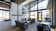 This contemporary lounge space makes an immediate impression with its dramatic double-height ceilings and expansive floor-to-ceiling window walls that flood the interior with abundant natural light and offer open views of the surrounding area. The architectural design blends industrial and modern elements, with exposed structural columns and a refined, polished aesthetic throughout.

At the heart of the space sits a sleek, linear fireplace framed in clean white paneling, serving as a striking focal point for the seating area. A carefully curated arrangement of patterned armchairs and accent seats surrounds a glass-topped coffee table, creating an inviting and cohesive gathering area. The furnishings are unified by a sophisticated blue and gold color palette, complemented by decorative throw pillows and accent pieces that add warmth and personality.

Abstract artwork in coordinating hues adorns the walls, reinforcing the space's contemporary character. Pendant lighting and track fixtures provide layered illumination, enhancing the ambiance during all hours. The neutral carpeted flooring grounds the space and adds a sense of comfort underfoot. Large glass entry doors connect the interior seamlessly to the outdoors, further amplifying the sense of openness and light that defines this well-appointed, single-floor space.