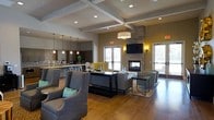 This well-appointed single-floor community space offers a sophisticated and welcoming atmosphere designed for both relaxation and social interaction. The main lounge area is anchored by a striking floor-to-ceiling stone fireplace, which serves as a focal point for multiple carefully arranged seating groups. Plush gray upholstered armchairs and sofas, accented with teal throw pillows, are positioned around dark-finished coffee and side tables, creating an inviting and comfortable environment.

The ceiling features elegant coffered beam detailing, adding architectural depth and character to the space. Recessed lighting, along with stylish pendant lights and decorative sconces, provides warm, layered illumination throughout. Rich hardwood flooring flows seamlessly across the entire area, complementing the neutral yet modern color palette.

Adjacent to the lounge, a bar or café-style counter area is equipped with pendant lighting and what appears to be a kitchen or refreshment station, making the space versatile for a variety of gatherings. Large windows and glass doors allow ample natural light to fill the room while offering views of the exterior surroundings.

An accent wall featuring a textured, patterned wallcovering adds a decorative element that balances the room's contemporary stone and wood finishes. The overall ambiance is modern, comfortable, and thoughtfully designed.