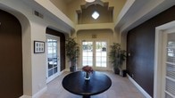 This sophisticated lobby makes a striking first impression with its grand, soaring ceiling that amplifies the sense of openness and arrival. A central decorative quatrefoil light fixture serves as a focal point overhead, drawing the eye upward and adding an architectural flourish to the space. The ceiling features structured geometric detailing, with layered molding that frames the upper walls and adds depth and dimension to the room.

The entrance is anchored by a set of elegant French doors that flood the interior with abundant natural light, offering views to the outdoors. Flanking the entry, tall potted bamboo plants introduce a natural, organic element that contrasts beautifully with the refined surroundings. A dark round pedestal table at the center of the room holds a floral arrangement, grounding the space with a welcoming touch.

The walls blend warm beige tones with rich dark brown accents, creating a refined and inviting atmosphere. Arched alcoves on either side of the entry add architectural character, while additional side doors with glass panes enhance connectivity and light flow. Light-toned tile flooring completes the polished aesthetic, making this lobby a well-appointed and welcoming gateway to the rest of the building.
