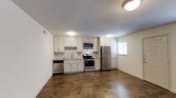 This single-floor space showcases a clean, modern design centered around an open-concept layout that seamlessly connects the kitchen and living areas. The kitchen is well-appointed with a full suite of stainless steel appliances, including a gas range with oven, over-the-range microwave, dishwasher, and a full-size refrigerator. White shaker-style cabinetry lines the walls, providing ample storage above and below the countertops, which feature a light-toned surface complemented by a white tile backsplash. A stainless steel sink is centrally positioned along the counter run, accompanied by a window that allows natural light to brighten the space. Dark hardwood-style flooring extends throughout, offering a warm contrast to the bright white cabinetry and neutral walls. Flush-mount ceiling lights provide consistent overhead illumination across the open floor plan. The layout allows the kitchen to flow naturally into the adjacent living area, creating a versatile and functional space. The overall aesthetic is contemporary and well-maintained, with a neutral palette that offers flexibility for various interior design preferences.