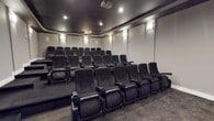 Located at Spring Creek, this dedicated screening room offers an immersive, cinema-style experience within a single-floor layout. The room is designed to replicate the feel of a professional movie theater, featuring multiple rows of tiered, stadium-style seating that ensures clear sightlines from every position. Each seat is a generously sized, cushioned reclining chair upholstered in dark fabric, complete with built-in armrests and cup holders for added comfort during extended viewings. The seating is arranged on a raised platform at the rear, providing an elevated perspective toward the front projection wall. Overhead, recessed ceiling lights offer adjustable ambient illumination, while wall-mounted sconce lighting along the sides adds a soft, atmospheric glow. A ceiling-mounted projector serves as the centerpiece of the entertainment setup, delivering high-quality visuals. The room's dark color palette—encompassing the charcoal ceiling, neutral walls, and patterned carpeted flooring—reinforces a focused, distraction-free viewing environment. The overall design balances functionality and comfort, making this space well-suited for private movie screenings, presentations, or group entertainment events.