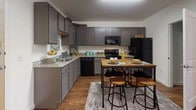 This studio is a compact yet thoughtfully arranged single-floor space that maximizes functionality through smart design choices. The open-concept layout connects the kitchen and living area seamlessly, creating an efficient flow throughout.

The kitchen is a standout feature, offering generous grey cabinetry that extends to the ceiling for maximum storage, granite-style countertops, and a full suite of black appliances including a refrigerator, range, dishwasher, and built-in microwave. Wood-look flooring runs throughout, adding warmth and visual continuity.

The main living area demonstrates clever use of space, utilizing an open shelving unit as a room divider to define a dedicated sleeping zone from the living and workspace areas. The sleeping area benefits from natural light through a window with drape panels, while the living side accommodates a sofa and small work desk comfortably.

The bathroom features a granite-topped vanity with an undermount sink, dark cabinetry, a large mirror, and tile flooring—offering a clean, polished finish. Overall, the studio presents a cohesive, neutral color palette accented with teal and yellow tones, creating a bright and inviting atmosphere throughout.