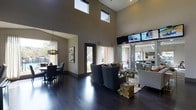 This stylish clubroom offers an inviting, upscale environment designed to accommodate a variety of social activities. The space is immediately distinguished by its dramatic double-height ceilings, which are punctuated by clerestory windows that draw in abundant natural light and create a sense of openness and grandeur. Dark-stained hardwood floors run throughout, providing a rich, polished foundation that anchors the room's contemporary aesthetic.

The layout is thoughtfully divided into functional zones. A comfortable lounge area features plush, modern armchairs arranged around a glass-topped coffee table with natural wood accents, complemented by a mounted flat-screen television for entertainment. An adjacent dining area provides ample seating around a sleek table beneath a statement pendant light fixture, making it well-suited for group gatherings or events.

Large windows dressed with floor-length curtains frame views of the surrounding exterior, further enhancing the bright and airy atmosphere. Neutral wall tones in warm taupe and gray serve as a refined backdrop, while carefully selected artwork adds character to the space. A glazed partition offers a visual connection to an adjoining area, reinforcing the open, community-focused design. Overall, the clubroom presents a polished, welcoming environment ideal for both casual relaxation and organized social events.