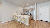 Located in Box Elder, South Dakota, this single-floor unit showcases a contemporary design highlighted by exposed industrial-style ductwork running through the living and bedroom areas, adding a distinctive architectural character throughout. The open-concept living area is bright and airy, featuring light wood-style flooring, recessed lighting, and a large sliding glass door that allows generous natural light to fill the space. The kitchen is well-equipped with stainless steel appliances, taupe cabinetry, a white quartz island with bar seating, and pendant lighting overhead, making it both functional and visually appealing. The living space flows naturally from the kitchen, creating an ideal environment for daily living and entertaining. The bedroom is a calm retreat with soft carpeting, a large window with blinds, and the same exposed ductwork detail carried throughout. A dedicated walk-in closet offers extensive wire shelving for organized storage. The bathroom features wood-style flooring, a tub-shower combination, a single vanity with white countertop, and matte black hardware accents. An additional utility area with an electrical panel and wire shelving provides practical storage and mechanical access. Overall, the space delivers a cohesive, modern aesthetic with warm neutral tones.