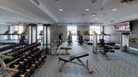 This fitness center offers a comprehensive workout environment thoughtfully designed to accommodate a wide range of exercise needs. The space is laid out across a single level and features a distinctive chevron-patterned carpet flooring that adds a modern aesthetic throughout. Recessed lighting and ceiling fans keep the atmosphere bright and comfortable, while large windows along one wall invite natural light and views of the surrounding streetscape.

The cardio zone is well-appointed with multiple Life Fitness treadmills, elliptical trainers, and a stationary bike, all positioned to face the windows — ideal for an engaging workout. A wall-mounted flat-screen television provides additional entertainment for those on the machines. The strength training area is equally impressive, featuring a multi-station cable machine, a full rack of dumbbells spanning a wide weight range, adjustable benches, and a cable crossover system. Stability balls and a medicine ball rack round out the functional fitness offerings.

Floor-to-ceiling mirrors line key sections of the space, enhancing the sense of openness and allowing users to monitor their form. The fitness center is accessible through glass-panel doors and includes clearly marked safety fixtures. Overall, the space is clean, modern, and fully equipped to support both cardiovascular conditioning and strength training routines.