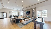 Located at 36 Stony Hill Road in Bethel, CT, this single-floor community space delivers a sophisticated, contemporary design across a generously sized layout. The living room serves as the centerpiece, showcasing warm wide-plank hardwood flooring, a striking slatted wood accent wall with an integrated linear fireplace, and a wall-mounted television flanked by marble-accented built-in shelving. A modern chandelier with sculptural pendants anchors the seating area, which is complemented by a shuffleboard table and ample natural light from large windows. Adjacent to the living area, a bar-style kitchen features deep navy cabinetry, a white stone-topped island with seating, and amber pendant lighting—offering a casual gathering spot with clean, upscale finishes. The dining room is enclosed behind sleek black-framed glass sliding doors, creating a semi-private meeting space. Inside, bold teal walls are adorned with illuminated wood-niche shelving, and a solid wood conference table is surrounded by modern swivel chairs. A transitional corridor with rounded gold-framed mirrors connects the spaces, reinforcing the cohesive, design-forward aesthetic throughout. Recessed lighting and abundant windows ensure the entire space feels bright, open, and welcoming.