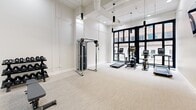 Located at 31 Pratt St in downtown Hartford, CT, this fitness center offers a well-appointed exercise space designed for a comprehensive workout experience. The room features soaring ceilings with exposed ductwork and sleek pendant lighting, lending an industrial-modern aesthetic to the bright, white interior. Large, floor-to-ceiling black-framed windows along one wall flood the space with natural light and provide views of the surrounding urban streetscape.

The fitness floor is finished with neutral, textured carpet throughout and is equipped with a thoughtfully curated selection of equipment. Cardio machines — including treadmills and stationary bikes — are positioned near the windows, while a cable resistance machine and adjustable weight bench occupy the central floor area. A multi-tier dumbbell rack with a full range of free weights is available for strength training. Wall-mounted mirrors further enhance the sense of openness and allow users to monitor their form.

Additional amenities include a wall-mounted flat-screen TV, a dual-level water fountain, and convenient access through a distinctive green-framed glass door. The clean, minimalist design and well-lit environment create a professional and motivating atmosphere suited for a variety of fitness routines.