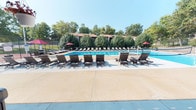 Located at 12405 Glenmeade Ct in Maryland Heights, Missouri, this outdoor pool facility offers a well-appointed recreational retreat designed for relaxation and leisure. The centerpiece is a large, rectangular swimming pool with clear blue water, framed by a generous concrete sundeck that provides ample space for sunbathing and socializing. Numerous cushioned lounge chairs are arranged across the deck, complemented by vibrant red market umbrellas that offer shaded respite on sunny days. Lush mature trees line the perimeter of the property, creating a natural privacy screen and a pleasant, park-like atmosphere. A covered pavilion area serves as the primary entry point to the pool deck, featuring floor-to-ceiling glass sliding doors with a keypad access system, ensuring a secure and controlled environment. The pavilion's open-air design transitions seamlessly between the interior passage and the outdoor pool area, with a staircase leading to upper-level access. Signage indicates the pool area is monitored by 24-hour electronic surveillance. A tennis or sport court is also visible in the background, adding to the range of outdoor amenities available at this community recreational space.