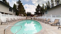 This inviting outdoor recreational facility centers around a generously sized, kidney-shaped swimming pool with clear turquoise water, framed by a broad, natural stone-tiled patio. The pool deck is well-equipped with numerous lounge chairs arranged for sunbathing, offering ample space for relaxation and leisure. A shaded umbrella seating area provides a comfortable retreat from the sun. The standout architectural feature of the complex is the pool house, constructed with distinctive warm-toned brick columns supporting a covered green-roofed overhang. The covered patio adjacent to the pool house is outfitted with outdoor dining furniture, making it an ideal gathering spot. Stone-tiled flooring extends throughout the patio area, lending a cohesive and refined aesthetic. White vinyl lattice fencing encloses the perimeter, providing privacy while maintaining an open, airy feel. The surrounding landscape is lush with tall evergreen and deciduous trees, creating a serene, natural backdrop. Adjacent residential buildings with neutral grey siding and white trim complement the setting. Pool rules are posted prominently at the pool house entrance, reflecting a well-maintained and organized recreational environment overall.