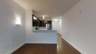 This single-floor space showcases a thoughtfully designed open-concept layout that seamlessly connects the kitchen and living areas. The kitchen is defined by rich, dark-toned cabinetry paired with stainless steel appliances, including a full-sized refrigerator, creating a sleek and modern aesthetic. A raised peninsula serves as a natural divider between the kitchen and the main living area, offering additional counter space and the potential for casual seating. Track lighting above the kitchen adds focused illumination, complementing the flush-mount fixtures visible in the adjoining hallway. The living area is characterized by wide-plank hardwood flooring in a deep, warm tone that extends throughout the space, adding a sense of cohesion and elegance. Crisp white walls and white baseboards create a clean, neutral backdrop that enhances the brightness of the interior. A hallway beyond the kitchen leads to additional areas of the unit, suggesting a well-organized floor plan. The overall ambiance is contemporary and move-in ready, combining functional design with modern finishes that appeal to a range of lifestyles.