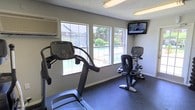 This single-floor fitness room is designed to provide a functional and comfortable workout environment. The space is outfitted with essential cardio equipment, including a treadmill and a recumbent exercise bike, both featuring individual digital consoles for tracking performance metrics. A wall-mounted flat-screen television is positioned prominently in the corner, allowing users to stay entertained during their workouts. The room is well-lit by overhead fluorescent lighting, supplemented by natural light streaming through multiple windows along one wall. These windows offer a pleasant view of an outdoor pool area surrounded by greenery and fencing, adding a refreshing visual element to the exercise experience. The flooring consists of durable rubber matting, which provides both safety and support during physical activity. A wall-mounted HVAC unit ensures climate control within the space, keeping it comfortable year-round. The neutral gray walls and practical layout contribute to a clean, utilitarian ambiance focused on fitness functionality. Overall, this fitness room serves as a convenient and well-appointed space for cardio-focused exercise.