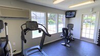 This single-floor fitness room is designed to provide a functional and comfortable workout environment. The space is outfitted with essential cardio equipment, including a treadmill and a recumbent exercise bike, both featuring individual digital consoles for tracking performance metrics. A wall-mounted flat-screen television is positioned prominently in the corner, allowing users to stay entertained during their workouts. The room is well-lit by overhead fluorescent lighting, supplemented by natural light streaming through multiple windows along one wall. These windows offer a pleasant view of an outdoor pool area surrounded by greenery and fencing, adding a refreshing visual element to the exercise experience. The flooring consists of durable rubber matting, which provides both safety and support during physical activity. A wall-mounted HVAC unit ensures climate control within the space, keeping it comfortable year-round. The neutral gray walls and practical layout contribute to a clean, utilitarian ambiance focused on fitness functionality. Overall, this fitness room serves as a convenient and well-appointed space for cardio-focused exercise.