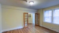 This single-floor space combines classic architectural charm with practical design. The main room is finished with warm, light-toned hardwood flooring that extends throughout, lending a clean and inviting feel to the interior. A standout feature is the pair of wood-framed French doors with glass pane inserts, which add character and elegance while allowing light to pass between spaces. These doors open to reveal a built-in closet with a dedicated shelving rod, providing organized storage. Adjacent to the closet, a secondary doorway leads to an additional room or utility space, enhancing the overall functionality of the layout. Natural light enters through a window fitted with blinds, brightening the room with a soft, diffused glow. The walls are finished in a neutral, light tone that complements the woodwork and contributes to the airy ambiance. White painted baseboards frame the perimeter of the room, adding a crisp, traditional detail. Ceiling-mounted lighting provides consistent illumination throughout. Overall, the space offers a straightforward, well-appointed interior with timeless design elements suited for comfortable everyday living.
