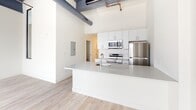 Located at 2501 Oakford Street in Philadelphia's Point Breeze neighborhood, this single-floor loft-style unit blends industrial character with contemporary finishes throughout. The space is defined by its dramatically high ceilings, exposed ductwork, and painted structural beams overhead, lending an authentic converted-warehouse aesthetic that sets it apart from conventional residences.

The open-concept living and kitchen area serves as the centerpiece of the unit. The kitchen is outfitted with crisp white cabinetry, a subway tile backsplash, stainless steel appliances including a range, microwave, and refrigerator, and a generous island with a light gray countertop and integrated sink. Light-toned hardwood flooring runs throughout, complementing the bright white walls and contributing to the airy feel of the space.

Large, multi-pane factory-style windows with black frames line the perimeter, flooding the interior with natural light and offering open views of the surrounding neighborhood. Roller shades provide adjustable privacy without compromising the industrial character of the windows.

A hallway connects the main living area to additional rooms, each accessible via barn-style hardware doors. The overall ambiance is clean, modern, and bright, with the exposed mechanical elements providing a distinctive urban edge that defines the loft's unique character.