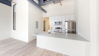 Located at 2501 Oakford Street in Philadelphia's Point Breeze neighborhood, this single-floor loft-style unit blends industrial character with contemporary finishes throughout. The space is defined by its dramatically high ceilings, exposed ductwork, and painted structural beams overhead, lending an authentic converted-warehouse aesthetic that sets it apart from conventional residences.

The open-concept living and kitchen area serves as the centerpiece of the unit. The kitchen is outfitted with crisp white cabinetry, a subway tile backsplash, stainless steel appliances including a range, microwave, and refrigerator, and a generous island with a light gray countertop and integrated sink. Light-toned hardwood flooring runs throughout, complementing the bright white walls and contributing to the airy feel of the space.

Large, multi-pane factory-style windows with black frames line the perimeter, flooding the interior with natural light and offering open views of the surrounding neighborhood. Roller shades provide adjustable privacy without compromising the industrial character of the windows.

A hallway connects the main living area to additional rooms, each accessible via barn-style hardware doors. The overall ambiance is clean, modern, and bright, with the exposed mechanical elements providing a distinctive urban edge that defines the loft's unique character.