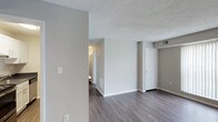 This single-floor residence at Crater Square offers a straightforward and functional layout finished with contemporary details throughout. The main living area is characterized by wide-plank laminate flooring in a cool grey-brown tone, which provides a modern aesthetic while offering durability. Neutral grey walls create a versatile backdrop suitable for a range of furnishing styles. The textured popcorn ceiling adds a layer of dimension to the space while housing a visible HVAC vent, indicating a central climate control system. A partial wall divides the main living space, subtly defining zones within the open layout. A white door leads to what appears to be a closet or storage area, contributing to the unit's practical storage options. A hallway visible in the background connects to additional areas of the unit, illuminated by a wall-mounted light fixture. White baseboards and trim throughout offer a clean contrast to the flooring and walls. The overall ambiance is understated and move-in ready, with a neutral palette that provides a blank canvas for personalization.