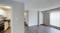 This single-floor residence at Crater Square offers a straightforward and functional layout finished with contemporary details throughout. The main living area is characterized by wide-plank laminate flooring in a cool grey-brown tone, which provides a modern aesthetic while offering durability. Neutral grey walls create a versatile backdrop suitable for a range of furnishing styles. The textured popcorn ceiling adds a layer of dimension to the space while housing a visible HVAC vent, indicating a central climate control system. A partial wall divides the main living space, subtly defining zones within the open layout. A white door leads to what appears to be a closet or storage area, contributing to the unit's practical storage options. A hallway visible in the background connects to additional areas of the unit, illuminated by a wall-mounted light fixture. White baseboards and trim throughout offer a clean contrast to the flooring and walls. The overall ambiance is understated and move-in ready, with a neutral palette that provides a blank canvas for personalization.