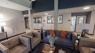 This single-floor commercial space presents a striking lobby or reception area defined by its dramatic double-height ceiling, which draws the eye upward to an open mezzanine level outfitted with cable railings and warm accent lighting. Structural columns finished in deep navy blue anchor the space visually, complementing the neutral gray and white walls and drop-tile ceiling below.

The seating area is thoughtfully appointed with a charcoal gray sofa accented by rust-orange throw pillows, paired with tufted gray leather armchairs arranged around a wood-framed coffee table. Decorative elements—including a tall table lamp, botanical stems, and small accent pieces—add warmth and a refined, professional character.

A row of interior-facing windows with dark frames lines the back wall, offering views into adjacent offices or workspaces, suggesting a functional professional environment such as a medical, legal, or corporate office suite. Corridor access is visible on both sides of the lobby, indicating multiple rooms or suites extending throughout the floor. The overall design balances modern aesthetics with a welcoming ambiance, conveying both professionalism and comfort.