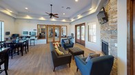 This single-story space is thoughtfully designed to accommodate both relaxation and gathering, combining modern finishes with warm, inviting details throughout. The main lounge area serves as the centerpiece, featuring tray ceilings with recessed lighting and a ceiling fan that enhance the sense of height and airflow. A floor-to-ceiling stacked stone fireplace anchors one wall, topped with a wall-mounted flat-screen television, creating a natural focal point. Mid-century modern seating in teal and charcoal is arranged around a tufted ottoman, promoting comfortable conversation. Rich wood-framed French doors open directly to an outdoor patio area, flooding the lounge with natural light and providing seamless indoor-outdoor access. Adjacent to the lounge, a dedicated dining area is lined with large multi-pane windows offering expansive views of lush landscaping and palm trees. Two glass-top dining tables with dark wood chairs take advantage of the natural light and scenic backdrop. The kitchen area includes warm wood cabinetry, granite countertops, tile flooring, and black appliances, offering a fully functional preparation space. Throughout, wood-look flooring and neutral wall tones create a cohesive, welcoming ambiance suited for a shared community setting.