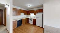 This single-floor unit offers a practical and efficient layout, making smart use of its available space. The open living area is carpeted and anchored by a large sliding glass door dressed with vertical blinds, allowing generous natural light to fill the room. A wall-mounted air conditioning unit and baseboard heating provide year-round climate control, while the textured ceiling adds a classic touch to the space.

The kitchen is well-appointed with warm wood cabinetry lining the walls, offering ample upper and lower storage. It features a full suite of white appliances, including a refrigerator, electric range, and a black dishwasher, all set on durable wood-look flooring. A flush-mounted ceiling light provides bright, even illumination throughout the kitchen.

The hallway connects the main living areas and includes a large sliding-door closet fitted with built-in shelving and a hanging rod, providing excellent storage capacity. The transition from wood-look flooring in the kitchen to carpet in the living and sleeping areas is clearly defined here.

The bathroom is clean and functional, featuring a full tiled tub-shower combination with white square tiles, a toilet, and mosaic tile flooring. The overall design of the unit is straightforward and practical, suited for efficient single-occupancy living.
