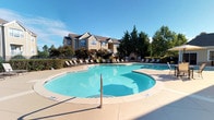 This well-appointed outdoor amenity area centers around a generously sized freeform swimming pool with a smooth concrete surround, offering ample space for lounging and recreation. The pool deck is lined with multiple reclining chairs positioned to take full advantage of the open sky and natural light throughout the day. A decorative stone fireplace anchors one end of the deck, serving as a functional and aesthetic focal point.

A pergola structure with stone-accented columns provides shaded seating areas equipped with high-top tables and chairs, ideal for casual outdoor dining or socializing. Ornamental container plantings with ornamental grasses add color and texture throughout the patio. An umbrella-covered seating arrangement offers additional sheltered options for outdoor relaxation.

The surrounding landscape features mature trees and manicured shrubs that provide privacy and a natural buffer. A wrought-iron perimeter fence defines the space while maintaining open sightlines to the greenery beyond. The adjacent clubhouse building, constructed in a classic craftsman-inspired style with neutral siding and a pitched roof, anchors the overall setting and contributes to the cohesive, resort-like ambiance of the outdoor space.