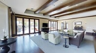 This single-story space showcases a refined interior defined by striking exposed dark wood ceiling beams set against a crisp, light-toned ceiling with recessed lighting. The open layout is generously proportioned, creating an airy and welcoming atmosphere throughout. Dark hardwood-style flooring anchors the space, complemented by a large area rug that defines the central seating arrangement, which features soft sage-green upholstered armchairs and additional neutral lounge seating with accent tables.

Multiple sets of full-height glass French doors line two walls, flooding the interior with abundant natural light and offering direct views and access to an outdoor patio area with visible landscaping. The seamless indoor-outdoor connection is a standout feature of the design. An arched architectural detail near the fireplace adds a classic Mediterranean or Southwestern design influence, harmonizing with the warm wood tones and neutral palette throughout.

A wall-mounted television serves as a functional focal point along one wall, while the overall furnishing arrangement encourages comfortable social gatherings. The combination of architectural detailing, natural light, and cohesive design elements gives this space a polished yet relaxed ambiance.