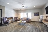 This single-floor space functions as a polished leasing or welcome center, thoughtfully designed to showcase available residences while providing a comfortable and inviting environment for visitors. The layout flows naturally from a well-appointed entry area into distinct functional zones, each finished with consistent patterned carpet flooring and neutral gray walls that lend the space a modern, cohesive aesthetic.

The entry zone features recessed lighting, a dark navy credenza with a round mirror above, and a frosted-glass interior door leading toward a secondary workspace or kitchenette area visible in the background. An exit door with glass panels allows natural light to filter into this transitional space.

A presentation area hosts a wall-mounted flat-screen television alongside display easels showcasing floor plan renderings, making it an ideal space for consultations. A ceiling fan, recessed lighting, and a window contribute to the comfortable, well-lit atmosphere.

The lounge area is a standout feature, centered around a sleek linear fireplace set into large-format tile cladding. A leather sofa, egg-style accent chair, arc floor lamp, and a white coffee table complete the seating arrangement. Large windows frame views of an outdoor pool and surrounding community, filling the space with natural light and creating a seamless indoor-outdoor connection.