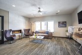 This single-floor space functions as a polished leasing or welcome center, thoughtfully designed to showcase available residences while providing a comfortable and inviting environment for visitors. The layout flows naturally from a well-appointed entry area into distinct functional zones, each finished with consistent patterned carpet flooring and neutral gray walls that lend the space a modern, cohesive aesthetic.

The entry zone features recessed lighting, a dark navy credenza with a round mirror above, and a frosted-glass interior door leading toward a secondary workspace or kitchenette area visible in the background. An exit door with glass panels allows natural light to filter into this transitional space.

A presentation area hosts a wall-mounted flat-screen television alongside display easels showcasing floor plan renderings, making it an ideal space for consultations. A ceiling fan, recessed lighting, and a window contribute to the comfortable, well-lit atmosphere.

The lounge area is a standout feature, centered around a sleek linear fireplace set into large-format tile cladding. A leather sofa, egg-style accent chair, arc floor lamp, and a white coffee table complete the seating arrangement. Large windows frame views of an outdoor pool and surrounding community, filling the space with natural light and creating a seamless indoor-outdoor connection.