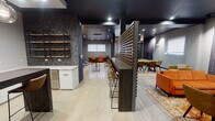Located at 5151–5163 N Sheridan Rd in Chicago's Edgewater neighborhood, this single-floor communal space blends industrial and contemporary design elements into a cohesive, multi-functional lounge environment. Exposed brick walls contrast with dark, textured wallcovering featuring a gold grid motif, creating a rich visual backdrop throughout. Dark ceilings with recessed lighting lend a moody, refined atmosphere across all zones.

The space is organized into several distinct areas. A lounge zone features high-back upholstered pod chairs in muted orange and slate tones, paired with ottomans and side tables, ideal for focused or semi-private conversations. Warm brass wall sconces illuminate the exposed brick accent wall nearby. A secondary seating area accommodates softer lounge furniture, including an orange sofa and tufted chairs arranged on a patterned area rug.

A slatted wood partition divides the main lounge from a bar-height communal workspace with leather-style barstools. Adjacent to this is a well-appointed kitchenette featuring white cabinetry, quartz countertops, a stainless steel sink, dishwasher, and open pipe-and-wood industrial shelving. A dark wood island with pendant globe lighting anchors the kitchen area. Polished concrete-style flooring ties the entire space together.