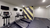 This fitness center is designed with both functionality and visual impact in mind. The most striking design element is the bold diagonal striping pattern adorning multiple accent walls, rendered in black and gold, which gives the space a dynamic, energetic atmosphere. The remaining walls are finished in clean white, providing contrast and keeping the overall environment bright despite the absence of natural windows.

The flooring throughout consists of dark, patterned carpet tiles that provide cushioning underfoot and contribute to the room's industrial-modern aesthetic. Exposed silver ductwork runs along the upper portion of one wall, adding an intentional industrial design touch.

The space is equipped with a comprehensive selection of cardio machines, including multiple treadmills, a stationary bike, and an elliptical trainer, all positioned to allow comfortable use. A dedicated free-weight area features a full rack of dumbbells in varying weights alongside adjustable workout benches. Wall-mounted televisions are positioned throughout for entertainment during workouts. A large mirror along one wall visually expands the space and allows users to monitor their form.

Recessed lighting provides consistent illumination across the entire fitness floor, and a water station near the entrance ensures convenient hydration access.