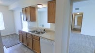 This two-story residence offers a practical and comfortable layout across its floors. The kitchen is equipped with warm-toned oak cabinetry spanning both upper and lower sections, providing generous storage space. A double stainless steel sink is set into a light-toned laminate countertop, accompanied by a white dishwasher beneath. A notable architectural feature is the pass-through window opening in the kitchen, which connects to the adjacent living area, allowing natural interaction between spaces and promoting an open feel. The tile flooring in the kitchen contrasts with the carpeted areas visible beyond, creating distinct zones within the home. A flush-mount ceiling light provides overhead illumination in the kitchen. The pass-through view into the living area reveals neutral-toned walls and carpeted flooring, suggesting a comfortable and cohesive interior palette throughout. The staircase visible in the background hints at the home's two-floor configuration, with the upper level accessible from the main living space. Overall, the residence presents a clean, functional aesthetic with classic wood finishes and a straightforward, well-organized floor plan.