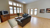 This well-appointed single-floor space offers an open, airy atmosphere defined by thoughtful design and abundant natural light. The main living area is a standout feature, boasting soaring ceilings that enhance the sense of openness and volume. A generous bank of large windows along one wall, framed in warm wood trim, floods the room with natural light and provides views of the surrounding exterior. Additional tall, narrow windows along the adjacent wall further brighten the space and reinforce the open feel.

The flooring throughout is light-toned hardwood, adding warmth and a clean, contemporary aesthetic to the interior. Neutral beige walls complement the natural wood accents found in the window trim and baseboards, creating a cohesive and welcoming palette. The living area is furnished with a substantial dark leather sofa and loveseat arrangement centered around a classic wooden coffee table, all anchored by a textured area rug. A potted plant adds a touch of greenery and life to the room.

Overhead, flush-mounted fluorescent lighting provides supplemental illumination, ensuring the space remains well-lit regardless of the time of day. The overall ambiance is calm, functional, and inviting — well-suited for gathering, relaxing, or professional use.