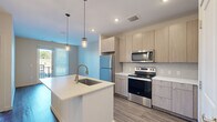 This thoughtfully designed single-floor residence offers a functional and modern layout across a compact yet efficient footprint. The open-concept kitchen serves as the centerpiece of the living area, featuring light wood cabinetry extending to the ceiling, sleek white quartz countertops, and a kitchen island with an undermount sink and gold-toned faucet. Stainless steel appliances — including a refrigerator, electric range, and over-the-range microwave — complement the contemporary aesthetic. Pendant lighting with brass-finished fixtures adds a warm, stylish touch above the island, while recessed lighting illuminates the space throughout. Wood-look plank flooring extends across the main living and kitchen areas, providing a cohesive and easy-to-maintain surface. A stacked washer and dryer unit is conveniently integrated into the layout, adding valuable in-unit functionality. The bedroom offers plush carpeting underfoot, a window with outdoor views, and access to a dedicated closet for storage. The bathroom is clean and neutral in design, featuring a full soaking tub with shower, a wood-toned vanity with white countertop, and wood-look flooring that ties into the rest of the home. Neutral wall tones throughout create a bright, move-in-ready atmosphere.