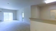 This single-floor residence offers a clean, open layout designed for comfortable everyday living. The main living area is finished with soft wall-to-wall carpeting in a neutral tone, providing a warm and quiet underfoot experience. The walls are painted in a crisp, light neutral shade throughout, contributing to a bright and airy atmosphere that is enhanced by natural light streaming in through multiple windows.

A defining architectural feature of the space is the built-in pass-through counter, which connects the kitchen area to the living space. This functional element adds visual interest while maintaining an open flow between areas. The counter features a recessed upper section, giving it a structured, purposeful design.

The living area is generously proportioned, with an open sightline that extends toward an adjacent room or hallway, suggesting a well-connected floor plan. The windows allow ample daylight to penetrate the interior, keeping the space feeling fresh and inviting. Electrical outlets and light switches are thoughtfully positioned along the walls for practical use.

Overall, this residence presents a move-in ready environment with a straightforward, functional design. Its neutral palette and open arrangement make it highly adaptable to a variety of furnishing styles and personal preferences.