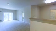 This single-floor residence offers a clean, open layout designed for comfortable everyday living. The main living area is finished with soft wall-to-wall carpeting in a neutral tone, providing a warm and quiet underfoot experience. The walls are painted in a crisp, light neutral shade throughout, contributing to a bright and airy atmosphere that is enhanced by natural light streaming in through multiple windows.

A defining architectural feature of the space is the built-in pass-through counter, which connects the kitchen area to the living space. This functional element adds visual interest while maintaining an open flow between areas. The counter features a recessed upper section, giving it a structured, purposeful design.

The living area is generously proportioned, with an open sightline that extends toward an adjacent room or hallway, suggesting a well-connected floor plan. The windows allow ample daylight to penetrate the interior, keeping the space feeling fresh and inviting. Electrical outlets and light switches are thoughtfully positioned along the walls for practical use.

Overall, this residence presents a move-in ready environment with a straightforward, functional design. Its neutral palette and open arrangement make it highly adaptable to a variety of furnishing styles and personal preferences.