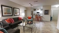 This single-floor space showcases an open-concept layout that seamlessly connects the living area to the kitchen, creating a cohesive and functional environment. The living room is anchored by a tufted dark charcoal sofa and a complementary teal accent chair, both adorned with vibrant coral-red throw pillows that add a striking pop of color against the neutral beige walls. A glass-and-metal coffee table sits atop a patterned area rug, enhancing the contemporary aesthetic. Track lighting runs along the ceiling, providing adjustable illumination throughout the space. Wall art in dark frames adds visual interest and a curated feel to the living area. The kitchen features white cabinetry, stainless steel appliances, and a peninsula counter with bold red bar stools, reinforcing the space's consistent color palette. Light wood-style flooring runs throughout, unifying the open floor plan and contributing to an airy, modern ambiance. The layout allows for clear sightlines from the living area to the kitchen, making the space feel both spacious and connected—ideal for everyday living and casual entertaining.