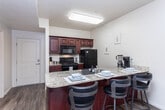 This single-floor residence offers a thoughtfully designed open-concept layout that seamlessly connects the kitchen, dining, and living areas. The kitchen is equipped with rich cherry-toned cabinetry, granite countertops, black appliances including a gas range, refrigerator, microwave, and dishwasher, as well as a breakfast bar with space for multiple bar stools — ideal for casual dining. Wood-look flooring runs throughout the main living areas, complementing the warm, neutral color palette. The living room is bright and airy, benefiting from multiple windows with vertical blinds that allow generous natural light. A ceiling fan adds comfort, and the space comfortably accommodates a sofa, coffee table, and entertainment setup. The residence includes two bedrooms, both carpeted and featuring large windows that bring in ample daylight. Each room offers a calm, restful atmosphere with clean lines and neutral tones. The bathroom features wood-look flooring, granite-topped cherry cabinetry, and a shower-tub combination. Throughout the unit, the design strikes a balance between functionality and modern comfort, making it a practical and inviting living space.