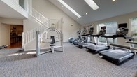 This fitness center offers a comprehensive workout environment designed to accommodate a wide range of exercise routines. The space is characterized by soaring vaulted ceilings with skylights that flood the interior with abundant natural light, creating an open and energizing atmosphere. Neutral-toned walls and patterned carpet flooring contribute to a clean, functional aesthetic throughout.

The main workout area is generously proportioned and organized into distinct training zones. A dedicated cardio section houses multiple treadmills, elliptical trainers, and stationary bikes, all positioned near large windows that provide views of the surrounding greenery. A cable machine and functional trainer system anchor the strength training zone, complemented by a medicine ball rack offering a variety of weights for dynamic exercises.

A separate free weights area includes a full dumbbell rack with a wide weight selection, an adjustable workout bench, and a wall-mounted fan for ventilation. French doors in this section open to an outdoor area framed by a stone wall, introducing fresh air and additional natural light. Large mirrors throughout enhance the sense of space and allow users to monitor their form. A staircase with white balusters is also visible, adding an architectural element to the overall design.