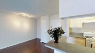 This single-floor residence offers a clean, open-concept design that seamlessly connects the kitchen and living areas. The kitchen is outfitted with white cabinetry that extends to the ceiling, providing ample storage, and features speckled granite countertops that lend a polished, functional aesthetic. A peninsula-style island extends into the space, creating a natural transition between the kitchen and the adjoining living area while offering additional prep and seating space. The flooring throughout is rich, dark-toned hardwood, adding warmth and contrast against the bright white walls and cabinetry. A textured ceiling adds subtle character to the interior, while a track lighting system installed along the ceiling ensures the living area is well-lit and adaptable. Neutral, light-toned walls throughout keep the space feeling airy and open. A single interior door provides access to an additional room or closet, contributing to the practical layout of the residence. Overall, the space presents a bright, well-maintained interior with a cohesive design palette that balances modern finishes with comfortable, everyday livability.