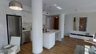 This single-floor unit showcases a contemporary open-concept layout that seamlessly blends the kitchen and living areas into one cohesive, functional space. The kitchen is outfitted with crisp white flat-panel cabinetry that extends to the upper walls, offering generous storage while maintaining a clean, minimalist aesthetic. A white quartz countertop island serves as the centerpiece of the space, providing additional prep area and casual seating. Stainless steel appliances, including a refrigerator and dishwasher, add a polished, modern touch, while a classic subway tile backsplash introduces subtle texture. Warm hardwood-style flooring runs throughout, creating an inviting contrast against the bright white palette. A structural column near the kitchen adds an architectural element that subtly defines the transition between cooking and living zones. Pendant lighting hangs above the island, complementing the recessed ceiling lights that ensure the space is well illuminated. The overall ambiance is bright, clean, and modern, making this a highly functional yet stylish environment suited for everyday living and entertaining alike.