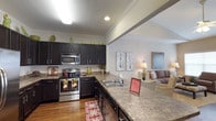 Located at 8254 County Road 64 in Daphne, Alabama, this single-floor residence offers a thoughtfully designed open-concept layout that seamlessly connects the kitchen and living areas. The kitchen is a standout feature, showcasing rich dark cabinetry that contrasts elegantly with granite-style countertops. Stainless steel appliances, including a range, microwave, and dishwasher, provide a modern and functional cooking environment. A double-basin sink sits along the extended counter, which opens directly to the living space, making it ideal for entertaining. Crown molding runs throughout, adding a refined architectural detail, while vaulted ceilings lend a sense of openness and volume to the space. Warm hardwood flooring flows through the kitchen and into the living area, creating a cohesive and inviting atmosphere. Recessed lighting and under-cabinet illumination ensure the kitchen is well-lit, while the adjacent living room benefits from soft ambient lighting. The living area is generously proportioned, offering comfortable room for furnishings. The overall design balances practicality with aesthetic appeal, making this a well-rounded and functional single-level home in the Daphne, Alabama area.
