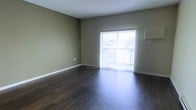 This single-floor space features a clean, functional layout with a focus on simplicity and natural light. The main living area is finished with rich, dark hardwood flooring that contrasts elegantly against the neutral beige-toned walls, creating a warm and grounded atmosphere throughout. White baseboards provide a crisp border between the walls and flooring, adding a polished finishing detail.

A prominent sliding glass door serves as the focal point of the living area, fitted with vertical blinds that allow for adjustable light control. The door opens directly onto a private balcony enclosed by a metal railing, offering views of the surrounding exterior environment and allowing natural light to fill the interior space during the day.

The walls are smooth and uniformly finished, providing a versatile backdrop suitable for various interior styles. Electrical outlets are positioned at standard intervals along the walls, and the layout appears open and unobstructed, maximizing usable floor space. The ceiling is flat and white, contributing to an overall sense of brightness and openness. The space presents a straightforward, move-in-ready environment with durable finishes well-suited for everyday living.
