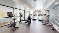 Located at 4333 N 6th Dr in Phoenix, AZ, this fitness center offers a thoughtfully designed, single-floor workout space. The layout is clean and open, providing ample room to move between a diverse selection of cardio equipment, including treadmills, elliptical trainers, stationary bikes, and a rowing machine. Large floor-to-ceiling windows line one side of the room, flooding the interior with natural light while offering views of the outdoor pool and surrounding courtyard. Full-length mirrored walls further enhance the sense of openness and allow users to monitor their form during workouts. The flooring consists of durable dark gray sport tiles suited for high-traffic fitness use. A standout design element is the bold geometric accent wall rendered in teal, cream, and navy, which adds a vibrant, modern character to the space. Recessed ceiling lighting ensures consistent, even illumination throughout. A wall-mounted flat-screen TV is positioned for easy viewing, and a hand sanitizer station is conveniently located near the entrance. Decorative indoor plants add a touch of greenery, softening the contemporary aesthetic. The overall ambiance is modern, energetic, and functional, making it an inviting environment for daily exercise.