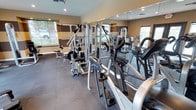 This fitness center offers a comprehensive workout environment designed to accommodate a range of exercise needs. The single-floor space is outfitted with a solid lineup of equipment, including multi-station cable machines, weight-stack strength units, elliptical trainers, and stationary bikes, providing options for both resistance training and cardiovascular workouts.

The room features neutral warm-toned walls accented by a contrasting horizontal band, contributing to a focused and energizing atmosphere. Large windows along the walls allow natural light to flow into the space, creating a bright and open feel. Recessed ceiling lighting supplements the natural light, ensuring consistent illumination throughout the room.

Full-length mirrors line one wall, visually expanding the space while allowing users to monitor their form during exercise. The flooring consists of durable, dark rubber-style tiles well-suited to heavy equipment and high-traffic use. The elevated ceiling height adds to the sense of spaciousness. Overall, the facility is thoughtfully arranged to maximize usable floor space while keeping equipment accessible and the environment comfortable for regular use.